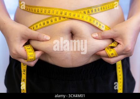 Primo piano di una donna che stringe il grasso addominale con entrambe le mani. Concetto di grasso corporeo, perdita di peso, fitness e stile di vita sano Foto Stock