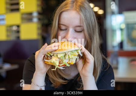 Una giovane donna siede a un tavolo con un grande hamburger ripieno di lattuga, pomodoro e altri condimenti, pronto a prendere un boccone in un vivace setti del ristorante Foto Stock