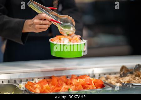 Primo piano delle mani di una donna con lunghe unghie rosse che usano pinze metalliche per servire il cibo in una caffetteria a pagamento. Indossa una giacca nera. Foto Stock