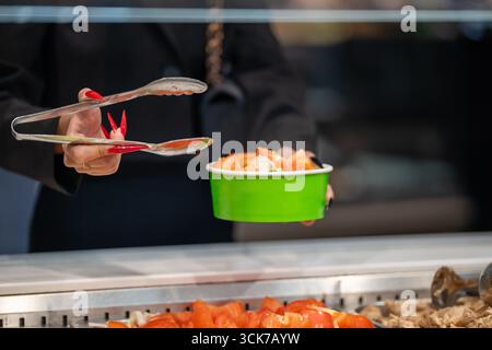 Primo piano delle mani di una donna con lunghe unghie rosse che usano pinze metalliche per servire il cibo in una caffetteria a pagamento. Indossa una giacca nera. Foto Stock