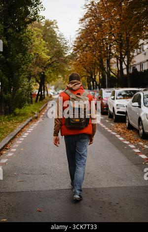 Uomo con zaino che cammina lungo la strada residenziale costeggiata da auto parcheggiate e alberi autunnali a Belgrado. Vista posteriore. Foto Stock