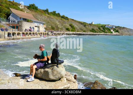 Folkestone Harbour, uomo e cane seduti accanto alla statua in bronzo della sirena Cornelia Parker, su una roccia a Sunny Sands Beach, Kent, Regno Unito Foto Stock