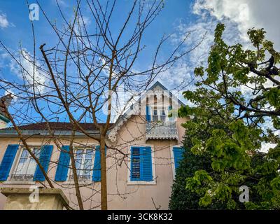 Affascinante casa francese con persiane blu brillante, balcone ornato e alberi da giardino sotto un cielo primaverile parzialmente nuvoloso. Foto Stock