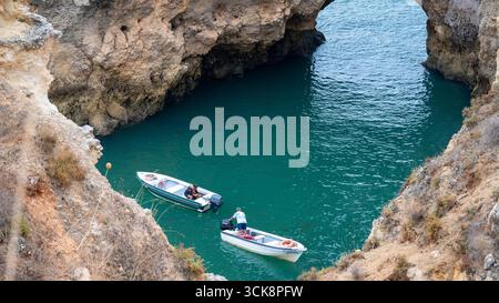 Piccole barche ancorate in una baia turchese a Ponta da Piedade, Lagos, Algarve, Portogallo, circondate da aspre scogliere e da un arco di roccia naturale sotto una o Foto Stock