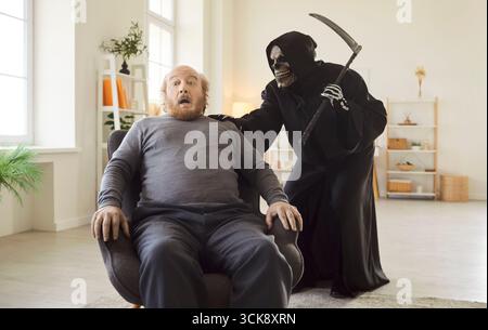 Uomo anziano spaventato seduto su una poltrona come un mietitore con un mantello nero che gli mette la mano sulla spalla. Foto Stock