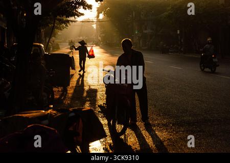 L'alba dorata illumina una strada vivace nel quartiere vecchio di Hanoi, Vietnam, con pedoni sagomati. Foto Stock