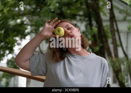 Donna giocosa con una maglietta grigia che tiene una mela verde all'occhio mentre fa un viso bacio in un giardino lussureggiante Foto Stock