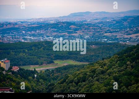 Un'ampia vista dalle colline di Termal che si affaccia su Yalova, Türkiye, mostrando fitte foreste in primo piano e aree urbane con montagne lontane in Foto Stock