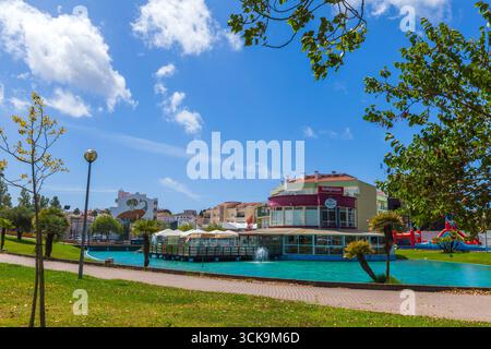 Un vivace parco urbano portoghese con un laghetto turchese, fontane, palme ed edifici moderni sotto un cielo blu brillante a Lourinhã Foto Stock