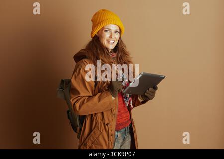 Una donna sorridente con un berretto giallo, una giacca marrone e una sciarpa a quadri tiene e punta su un tablet, pronta per viaggiare o avventure all'aperto con il suo backpa Foto Stock