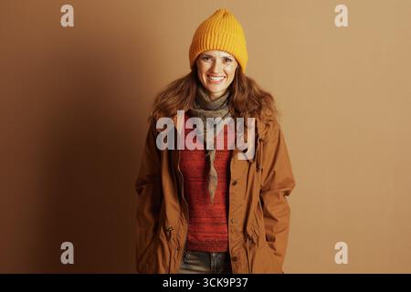 Una donna allegra con capelli marroni ondulati, vestita con un berretto in maglia gialla, un maglione ruggine e una giacca con cappuccio marrone, sorride alla fotocamera, trasudando calore e calore Foto Stock