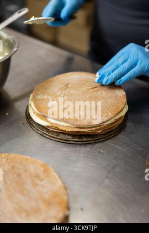 Chef che assembla la torta a strati con panna in cucina professionale, primi piani sulle mani e lavorazione Foto Stock