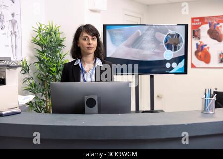 La receptionist femminile si trova vicino al computer al banco, aggiornando e mantenendo le cartelle cliniche dei pazienti. Ritratto del personale ospedaliero alla reception, guardando la macchina fotografica, pronto ad accogliere i visitatori della clinica. Foto Stock
