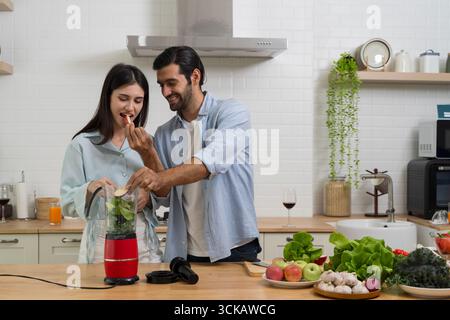 Coppia felice che prepara frullato in cucina moderna, uomo che dà da mangiare fette di mele alla donna, frutta e verdura fresca, stile di vita sano, nutrizione, amore, divertimento Foto Stock
