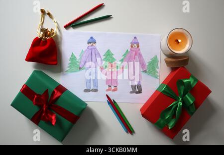 Disegno di una famiglia felice durante le passeggiate invernali, genitori e bambini che si tengono per mano. Le opere d'arte sono posizionate su una superficie bianca, circondate da regali, candele, astuccio, pe Foto Stock