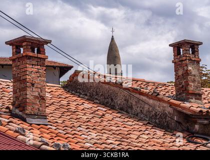 Stanjel, Slovenia - 27 luglio 2025: Vecchi camini in mattoni su un tetto piastrellato con una torre lontana. Foto Stock