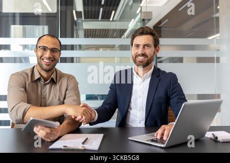 Due uomini d'affari sorridenti stringono la mano dopo aver raggiunto un accordo durante una riunione produttiva in un ufficio moderno, dimostrando il lavoro di squadra, la collaborazione e il successo della negoziazione Foto Stock