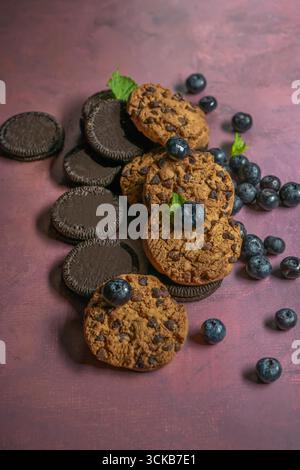 Una selezione di biscotti gourmet al cioccolato e ai mirtilli, un bicchiere di frullato di frutti di bosco con una paglia e mirtilli freschi sparsi su un luminoso e moderno Foto Stock