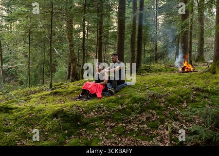 Due persone si godono un momento sereno insieme, uno che fa un massaggio all'altro, circondato da alberi alti, muschio morbido e un fuoco delicato che scoppia dentro Foto Stock