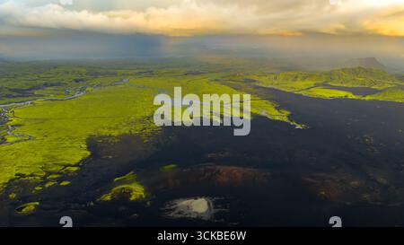 Immagine aerea del terreno ricoperto di muschio verde dell'Islanda, suolo vulcanico scuro, fiumi tortuosi e pianoro lontano sotto un cielo dorato con luce solare soffusa Foto Stock