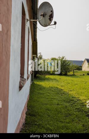 Una casa rurale in Bielorussia con pareti intonacate bianche, finiture rosa e una parabola satellitare. Un prato verde e cespugli incorniciano la scena, con un'altra casa vicina Foto Stock