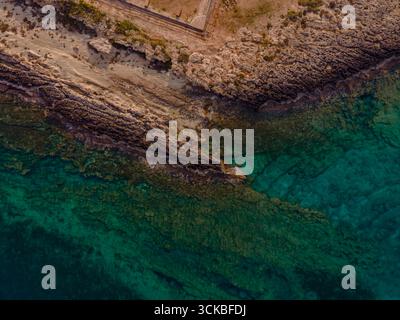 Immagine aerea della costa di Cipro che mostra scogliere rocciose, acque turchesi del Mediterraneo, macchie di sabbia dorata e una struttura artificiale lungo il bordo. Foto Stock