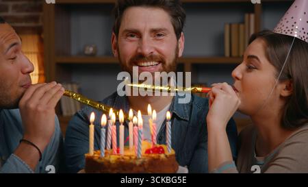 Buon compleanno adulto uomo caucasico con torta bruciare candele sorridente guardando macchina fotografica due multietnico ragazzo donna che soffia in festa fischietta a fischietti Foto Stock