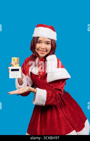 Giovane donna che indossa il costume di babbo natale, con un terminale pos con una carta di credito inserita, suggerendo shopping e spese natalizie Foto Stock