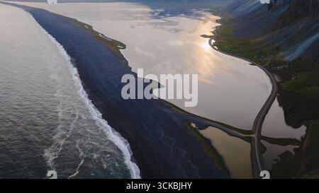 Una spettacolare vista aerea di una spiaggia di sabbia nera in Islanda, con l'oceano da un lato, una laguna riflettente dall'altro e montagne sullo sfondo Foto Stock