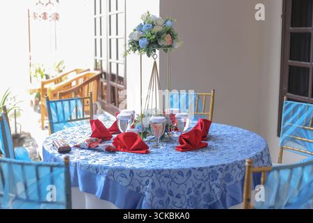 Un tavolo da pranzo splendidamente allestito con una tovaglia blu, tovaglioli rossi e un pezzo centrale floreale, circondato da eleganti sedie Foto Stock