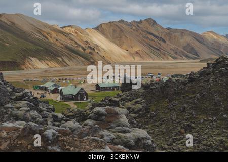 Campeggio nella regione islandese di Landmannalaugar con edifici dal tetto verde, tende e veicoli, circondato da rocce laviche muschiate e colline colorate. Foto Stock