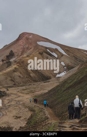 Un gruppo di escursionisti attraversa un aspro sentiero nella regione islandese di Landmannalaugar, con montagne riolite colorate e macchie di neve sotto un coperto Foto Stock