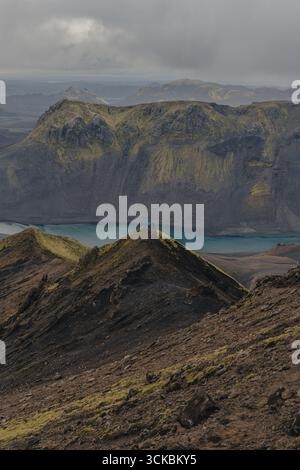 Un fiume turchese scorre sotto ripide scogliere coperte di muschio in Islanda. Una figura solitaria in una giacca blu sta su una cresta sotto un cielo coperto. Foto Stock