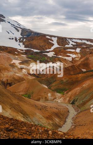 Vivaci montagne di riolite arancione, rosso e marrone con macchie di neve e un fiume tortuoso sotto un cielo coperto a Kerlingarfjoll, Islanda. Foto Stock