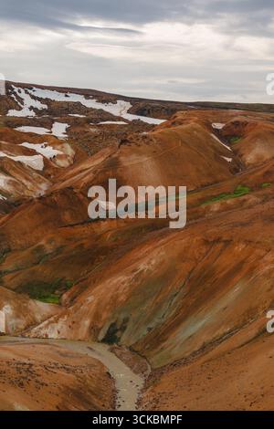 Vivaci colline riolite arancioni, rosse e marroni con macchie di neve sparse, un torrente tortuoso e un cielo coperto a Kerlingarfjoll, Islanda. Foto Stock