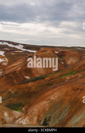 Vivaci colline riolite arancioni, rosse e marroni con macchie di neve sparse a Kerlingarfjoll, Islanda, sotto un cielo coperto con vegetazione sparsa. Foto Stock