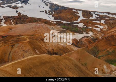 Vivaci montagne riolite a Landmannalaugar, Islanda, con sfumature arancio, rosso e marrone, macchie di neve, muschio verde e un picco innevato sotto le nuvole Foto Stock