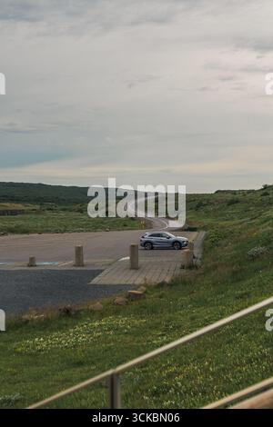 Un'auto argentata si trova in un piccolo lotto pavimentato circondato da erba verde e vegetazione bassa, con una strada tortuosa e un cielo nuvoloso nella campagna islandese. Foto Stock