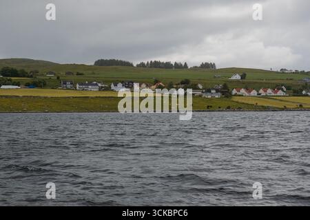 Tranquillo villaggio norvegese su una costa erbosa, caratterizzato da case con tetti rossi e bianchi, acque calme in primo piano e verdi colline sullo sfondo Foto Stock