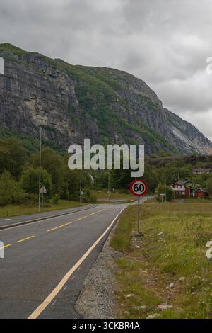 Una strada panoramica a Lysebotn, Norvegia, delimitata da campi erbosi e alberi, con scogliere rocciose, case rosse e bianche e un cielo coperto sullo sfondo Foto Stock