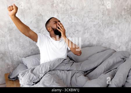 Uomo che sbadiglia e si allunga a letto durante la routine mattutina a casa Foto Stock