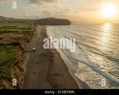 Vista aerea della luce dorata del sole che baciano le sabbie nere della spiaggia, dove le onde schiumose si infrangono contro la costa vicino alle scogliere, Black Beach, Azzorre, Portogallo. Foto Stock