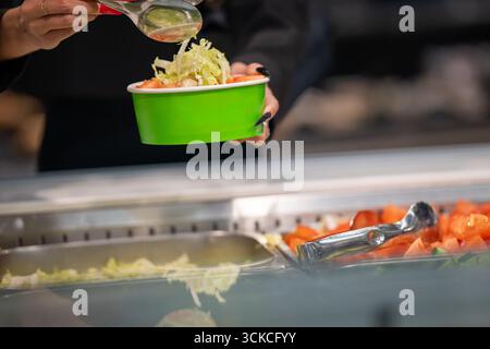 Primo piano delle mani di una donna con lunghe unghie rosse che usano pinze metalliche per servire il cibo in una caffetteria a pagamento. Indossa una giacca nera. Foto Stock
