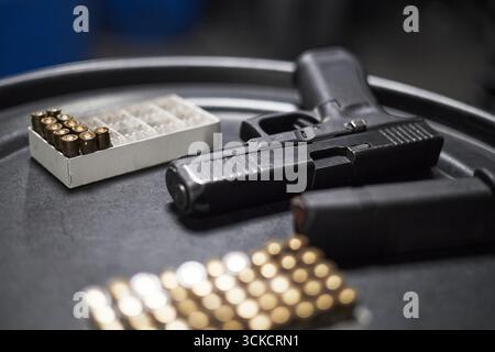 Pistola appoggiata su una superficie rotonda nera con caricatore carico nelle vicinanze, scatole di munizioni disposte in primo piano e sullo sfondo, fuoco su armi da fuoco e proiettili, nessuna persona visibile Foto Stock