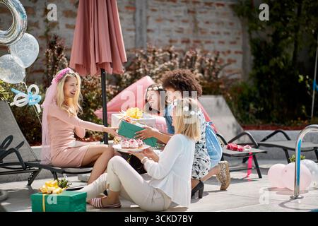 Gruppo di giovani donne multirazziali che si divertono accanto alla piscina, regalano regali alla bionda caucasica, ridendo Foto Stock
