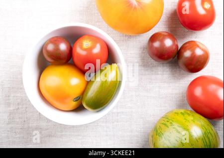 Pomodori freschi e maturi di varie varietà, colori e dimensioni, in una ciotola bianca, su tessuto di lino. Foto Stock