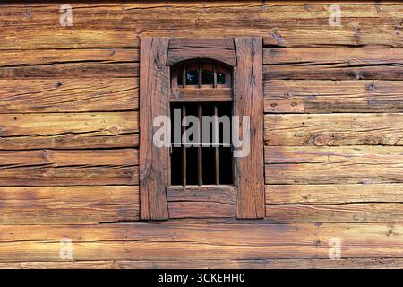 Facciata della cabina in legno con una minuscola finestra barrata inserita in tronchi orizzontali impilati, che mostrano nodi, granaglie ed età. Struttura robusta e simmetria f Foto Stock