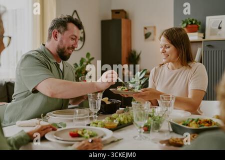 Giovane uomo caucasico che serve cibo alla giovane donna caucasica durante il pasto di famiglia Foto Stock