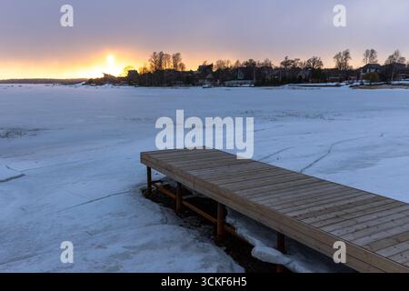 Tranquillo paesaggio invernale caratterizzato da un lago ghiacciato, un molo di legno che si estende verso la superficie ghiacciata e un tramonto incandescente che proietta una calda luce su dista Foto Stock