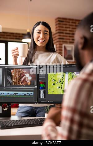 Una donna asiatica sorridente sta dietro i monitor del desktop mentre il ragazzo nero modifica le riprese video in casa. Una coppia felice e diversificata che condivide momenti di gioia lavorando insieme a progetti creativi. Foto Stock